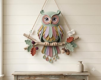 May include: A crocheted owl wall hanging in sage green, dusty pink, and mustard yellow, perched on a natural wooden branch. The owl features large eyes, layered wing feathers, and a long tail. Decorative crocheted oak leaves and acorns hang from the branch, which is suspended by a jute cord. Two small white tags are attached to the cord.
