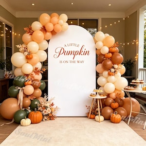 May include: A white arched backdrop with the text A LITTLE PUMPKIN IS ON THE WAY in orange, surrounded by a balloon garland in shades of cream, orange, brown, and olive green, accented with small pumpkins and dried floral arrangements.