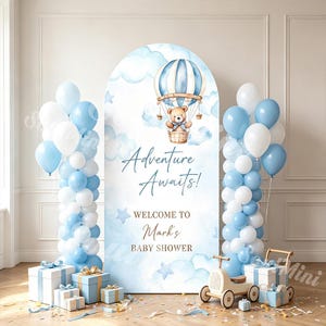 May include: An arched backdrop featuring a watercolour illustration of a teddy bear in a blue and white hot air balloon, with the text Adventure Awaits! Welcome to Mark's Baby Shower. Flanked by blue and white balloon columns and gift boxes.