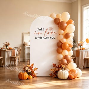 Op de afbeelding: Een witte boogvormige achtergrond met de tekst FALL in love WITH BABY AMY en een esdoornblad-afbeelding. Het is versierd met een tros crème, beige en oranje ballonnen, samen met oranje en witte pompoenen en herfstbladeren aan de voet.