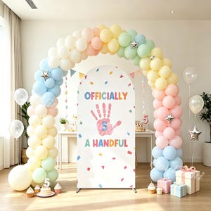 May include: A large arched party backdrop featuring the text "OFFICIALLY A HANDFUL" with a pink handprint graphic containing the number 5. The backdrop is framed by a colorful balloon arch in pastel shades of blue, yellow, pink, and green.