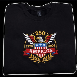 Könnte beinhalten: Ein schwarzes Sweatshirt mit Rundhalsausschnitt und einer zentralen Stickerei, die einen Weißkopfseeadler über einem Schild mit rot-weiß-blauen Streifen und drei Sternen zeigt. Die Zahlen 250, 1776, 2026 und das Wort AMERICA sind in Gold gestickt.