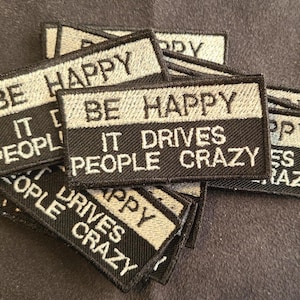 Könnte beinhalten: Eine Ansammlung rechteckiger bestickter Aufnäher mit zweifarbigem Design. Der obere Teil ist hellgrau mit dem schwarzen Text BE HAPPY, der untere Teil ist schwarz mit dem weißen Text IT DRIVES PEOPLE CRAZY in gestickten Buchstaben.