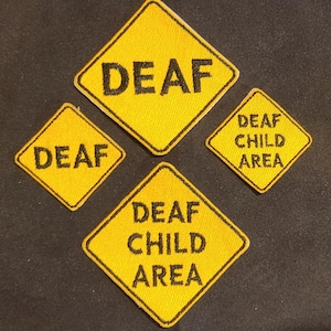 May include: Four diamond-shaped embroidered patches in bright yellow with black borders and black text. Two patches feature the word DEAF, while the other two patches display the text DEAF CHILD AREA in a stacked format on a textured fabric surface.