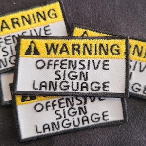 May include: A collection of rectangular embroidered patches featuring a yellow top bar with a black warning triangle icon and the text WARNING. Below this, a white section displays the black embroidered text OFFENSIVE SIGN LANGUAGE.