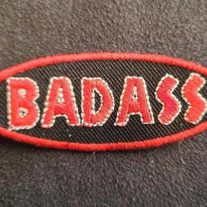May include: An oval-shaped embroidered patch featuring a black background with a red border. The word BADASS is stitched in red capital letters across the centre of the patch, with each letter outlined in a thin white thread for added definition.