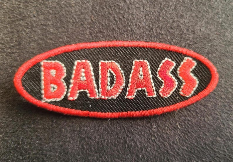 May include: An oval-shaped embroidered patch featuring a black background with a red border. The word BADASS is stitched in red capital letters across the centre of the patch, with each letter outlined in a thin white thread for added definition.
