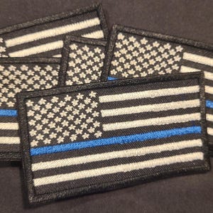 May include: A collection of rectangular embroidered patches featuring a stylized American flag design. Each patch displays a black background with white stars and stripes, accented by a single horizontal blue line running across the center.
