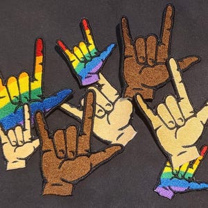 May include: A collection of embroidered patches featuring hand gestures in the I love you sign language configuration. The patches display various skin tones and rainbow-colored patterns, each outlined with a black border on a dark background.