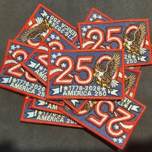 Op de afbeelding: Een verzameling rechthoekige geborduurde patches met een groot rood en wit getal 250, een Amerikaanse zeearend en de tekst 1776-2026 AMERICA 250. De patches hebben een blauwe achtergrond met witte sterren en een rode rand.