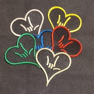May include: A collection of five heart-shaped embroidered patches, each featuring a black center with a contrasting colored border and a matching colored I love you hand sign gesture stitched in the middle. The borders are white, yellow, red, blue, and green.