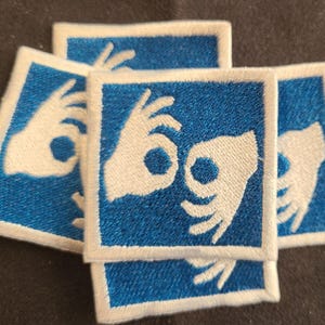 May include: A collection of square embroidered patches featuring a blue background and a white border. Each patch displays a white graphic of two hands forming the okay gesture, with the fingers and thumbs creating a circular shape in the center.