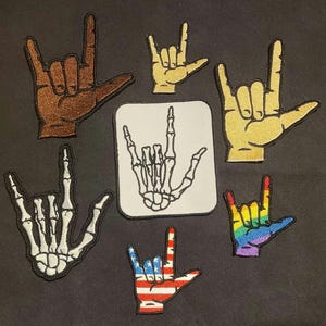 May include: A collection of seven embroidered patches featuring the American Sign Language I love you hand gesture. Designs include a brown hand, a yellow hand, a white skeleton hand, a rainbow-colored hand, and a hand with the American flag pattern.