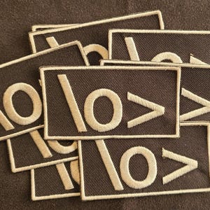 May include: A collection of rectangular embroidered patches featuring a dark brown background with a light beige stitched design consisting of a forward slash, a circle, and a greater-than symbol arranged horizontally across the center of each patch.