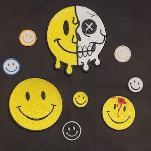 May include: A collection of embroidered patches featuring various smiley face designs. One large patch shows a yellow smiley face melting into a white skull with an X-shaped eye. Other patches include standard yellow, blue, and white smileys.