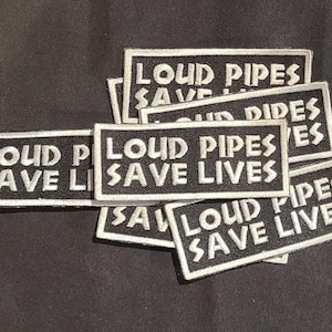 以下が含まれることがあります： 黒い背景に白い縁取り、そして太いブロック体の白い文字で「LOUD PIPES SAVE LIVES」と刺繍された長方形のワッペンが複数枚。ワッペンは重なり合うように積み重ねられています。