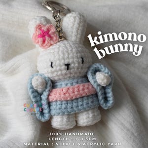 May include: A white crochet bunny keyring wearing a light blue kimono with a pink sash. The bunny features a pink flower on its ear, black embroidered eyes and nose, and a metal keyring. Text on the image reads: kimono bunny, 100% HANDMADE, LENGTH : +-8.5CM, MATERIAL : VELVET & ACRYLIC YARN.
