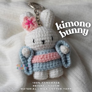 Può includere: Un portachiavi a forma di coniglio all'uncinetto bianco che indossa un kimono azzurro con una fascia rosa e un fiore rosa sull'orecchio. Il testo recita kimono bunny, 100% handmade, height: +-10cm, material: milk cotton yarn. Con anello.