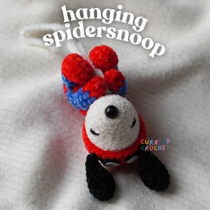 Puede incluir: Un juguete de peluche tejido a ganchillo con cabeza blanca, ojos y orejas negras, que lleva un traje rojo y azul con un lazo blanco para colgar. El texto "hanging spidersnoop" aparece arriba y "CURATED CROCHET" está impreso en letras pequeñas de colores.