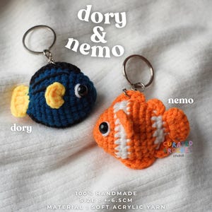 May include: Two crochet fish keychains labeled dory & nemo. The blue fish has yellow fins and black stripes, while the orange fish has white stripes. Both have black bead eyes and metal rings. Text includes 100% HANDMADE, SIZE : +-6.5CM, MATERIAL : SOFT ACRYLIC YARN.