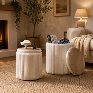 May include: Two cylindrical ottomans with vertical ribbed white fabric sides and removable round light wood lids. One ottoman is closed with a small white bowl and diffuser on top, while the other is open, revealing books and a patterned fabric inside.