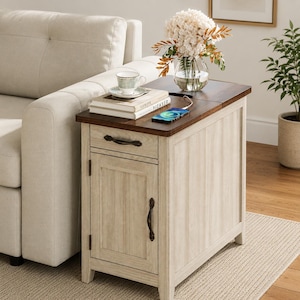 May include: A rectangular end table with a dark brown wooden top and a cream-colored base featuring vertical paneling. The table includes one drawer and one cabinet door with dark metal handles, a glass vase with flowers, books, and a smartphone.