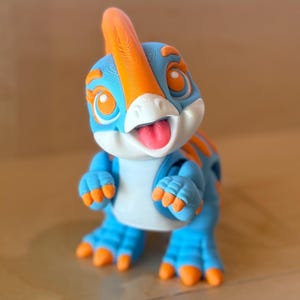 May include: A 3D printed dinosaur figurine featuring a blue body with bright orange accents on the head crest, eyes, back, and claws. The creature has a white snout, large orange eyes, and a small pink tongue sticking out of its open mouth.