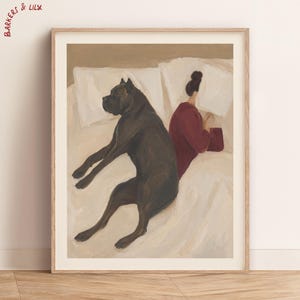 May include: A framed art print featuring a painting of a large black dog lying on white bed sheets next to a figure wearing a dark red long-sleeved top. The figure has dark hair in a bun and is facing away. The text BARKERS & LILY appears in red.