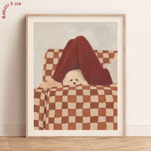 Pode incluir: Uma impressão artística emoldurada intitulada BARKERS & LILY com uma pintura de um pequeno cão branco espreitando por baixo de calças vermelho-escuras. O sujeito repousa num sofá com um padrão de xadrez castanho e bege, mostrando pés descalços.