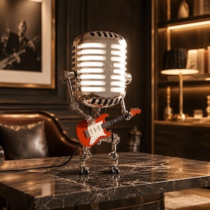 May include: A silver, vintage-style microphone lamp shaped like a robot figure. The microphone head glows with internal light. The robot has articulated metal limbs and is holding a small, bright orange electric guitar with a white pickguard.