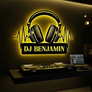 Puede incluir: Un letrero LED de pared con una silueta negra de auriculares centrada sobre una línea de latido y una placa rectangular con el texto DJ BENJAMIN. El letrero emite un brillo amarillo intenso que ilumina el entorno oscuro del estudio.