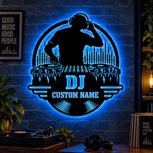 May include: A circular wall sign featuring a black silhouette of a DJ wearing headphones at a mixing console, illuminated by a bright blue LED light. The design includes a crowd, sound waves, and the text DJ CUSTOM NAME in bold lettering.