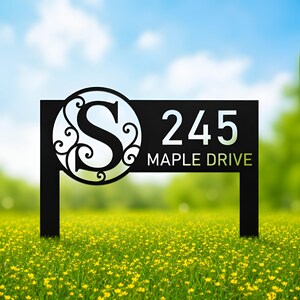 May include: A black metal address sign featuring a circular cutout with a decorative letter S, the number 245, and the text MAPLE DRIVE. The sign is supported by two vertical posts and displays a clean, high-contrast design for outdoor display.