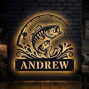 May include: A black metal wall sign featuring a backlit silhouette of a fish jumping out of water with a fishing rod arched above it. The rectangular base at the bottom contains the name ANDREW in bold, capitalised, cut-out lettering.
