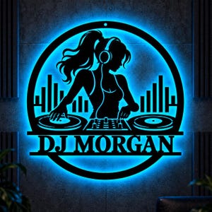 May include: A circular black metal wall sign featuring a silhouette of a DJ wearing headphones, positioned behind two turntables and a mixer. The design includes a graphic equalizer and the text DJ MORGAN, all illuminated by a bright blue LED light.
