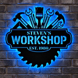 May include: A black metal wall sign with bright blue LED backlighting featuring a circular saw blade base, various hand tools like hammers and wrenches at the top, and the text STEVEN'S WORKSHOP EST. 1980 centered in a rectangular frame.