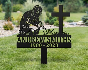 May include: A black metal garden stake memorial featuring a silhouette of a welder wearing a helmet and holding a welding torch. To the right is a cross above grass. The text reads ANDREW SMITHS and the dates 1980 - 2023 in a bold font.