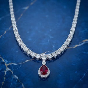 May include: A silver-toned necklace featuring a continuous row of round, clear, faceted stones. The center of the necklace displays a prominent pear-shaped, deep red stone surrounded by a halo of smaller, clear, round, faceted stones.