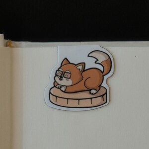 May include: A magnetic bookmark featuring a cartoon illustration of a brown fox wearing black-rimmed glasses. The fox is lying down on a light tan circular wooden log slice. The bookmark has a white border and a smooth, die-cut shape.