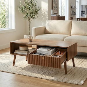 May include: A rectangular wooden coffee table with a walnut finish, featuring four tapered legs, an open side shelf, and a central pull-out drawer with a vertical ribbed front panel and a small circular gold knob, containing books and stationery.
