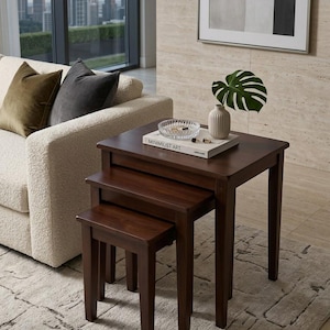 May include: A set of three dark brown wooden nesting tables with square tops and straight legs. The largest table holds a white book titled MINIMALIST ART, a clear glass bowl, and a small ribbed vase containing a single green monstera leaf.