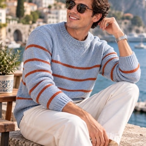 Op de afbeelding: Een lichtblauwe gebreide trui met horizontale roestkleurige strepen, een ronde hals en lange mouwen met geribbelde boorden. De trui wordt gecombineerd met een witte broek, wat zorgt voor een casual outfit voor buitenomgevingen.