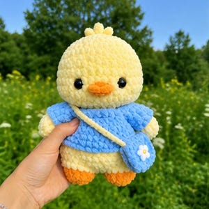 May include: A yellow crocheted chick plush toy wearing a blue shirt and a matching blue crossbody bag with a small white flower detail. The chick has black eyes and an orange beak, crafted from soft yarn with a textured, fuzzy appearance.