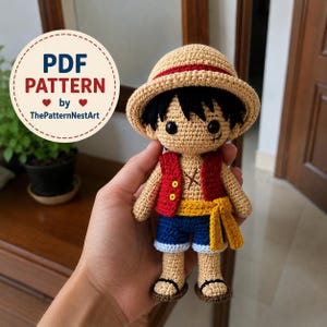 Puede incluir: Una muñeca de ganchillo con pelo negro, que lleva un sombrero de paja beige con una banda roja, un chaleco rojo con botones amarillos, pantalones cortos azules y sandalias. Incluye una X cosida en el pecho y el texto PDF PATTERN by ThePatternNestArt.