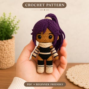 May include: A small amigurumi doll with dark brown skin, black eyes, and dark purple hair styled in a high ponytail. The doll wears a black and beige outfit with striped sleeves and a beige neck scarf. Text reads CROCHET PATTERN and PDF BEGINNER FRIENDLY.