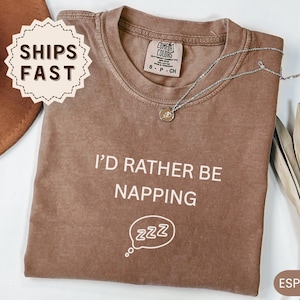 May include: A brown short-sleeved t-shirt with a crew neckline. The front features the white text I'D RATHER BE NAPPING above a white graphic of a speech bubble containing the letters ZZZ. The shirt has a Comfort Colors brand label at the neck.