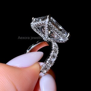 Op de afbeelding: Een zilverkleurige ring met een grote rechthoekige radiant-geslepen centrale steen omringd door een halo van kleine ronde steentjes. De ringband is ook bezet met kleine ronde steentjes aan de zijkanten. De tekst Aexora jewells is zichtbaar.