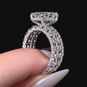 May include: A silver-toned ring featuring a large rectangular center stone set in a basket with pavé accents. The band is composed of three rows of round-cut stones, with the text Aexora Jewells visible on the side of the metal setting.
