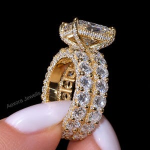 May include: A gold ring featuring a large rectangular radiant-cut centre stone set in a basket with small pavé diamonds. The band consists of two rows of round brilliant-cut stones set in gold. The text Aexora Jewells is visible on the side.