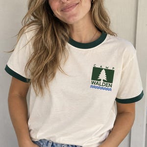 Può includere: Una t-shirt ringer color crema con bordi verde scuro sul colletto e sulle maniche. Il petto presenta una grafica con un quadrato verde scuro contenente un pino bianco, la scritta CAMP WALDEN e linee ondulate blu sottostanti.